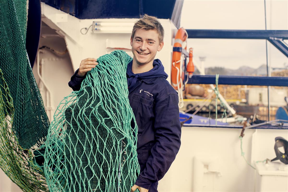 Mann i blå overall holder fiskegarn over skulderen. - Klikk for stort bilde